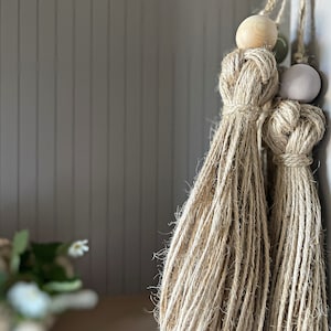Può includere: Due frange in macramé beige con perline di legno e grigio chiaro appese a un muro.