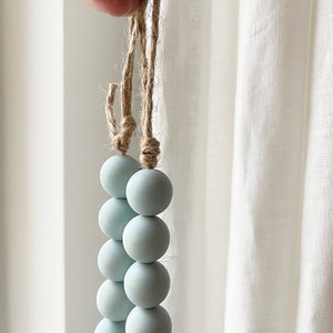 Puede incluir: Una guirnalda de cuentas de color azul claro con un colgador de cuerda natural. La guirnalda est&aacute; hecha de cuentas de madera y es perfecta para a&ntilde;adir un toque de color y textura a cualquier espacio.