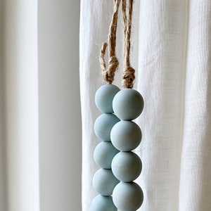 Puede incluir: Un conjunto de cuentas de madera de color azul claro enhebradas en un cord&oacute;n de cuerda natural. Las cuentas son redondas y lisas, y el cord&oacute;n est&aacute; anudado en la parte superior.