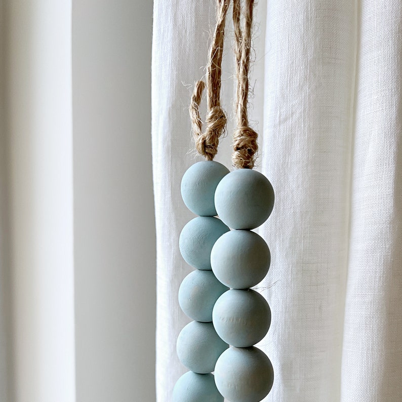 Puede incluir: Un conjunto de cuentas de madera de color azul claro enhebradas en un cord&oacute;n de cuerda natural. Las cuentas son redondas y lisas, y el cord&oacute;n est&aacute; anudado en la parte superior.
