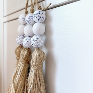 Könnte beinhalten: Eine weiße und blaue gemusterte Keramikperlenkette mit einem weißen und blauen Blumendesign. Die Girlande ist mit Naturbast gebunden und hat einen Quasten am Ende.