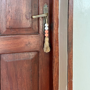 May include: A wooden door with a brass door handle and a tassel hanging from it. The tassel is made of beige fabric and has three beads: two peach-colored and one white.