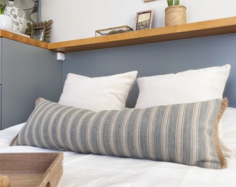 May include: A white bed with a blue and white striped lumbar pillow and two white pillows. The bed is against a blue wall with a wooden shelf above it.