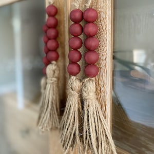 May include: Two burgundy wooden bead garlands with natural jute tassels. The garlands are hanging from a wooden surface.