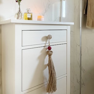 Peut inclure: Une commode blanche à trois tiroirs, chacun avec un bouton. Le tiroir supérieur a un pompon décoratif suspendu au bouton. Le pompon est fait de fibres naturelles et a une perle rouge au sommet.