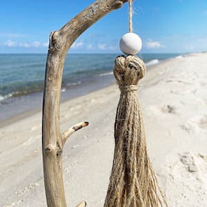Peut inclure: Ornement décoratif suspendu avec une branche de bois flotté naturelle. Une perle en bois blanc est suspendue à une corde de jute, au-dessus d'un long gland en jute. Le fond montre une plage de sable et un ciel bleu.