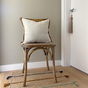 Puede incluir: Una silla de madera con un cojín de lino natural. La silla tiene un asiento tejido y un acabado marrón claro. La silla está sentada sobre una alfombra tejida con rayas de colores.
