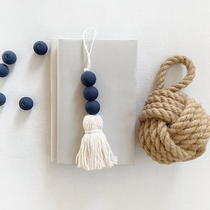 Könnte beinhalten: Ein marineblauer Quaste mit einem weißen Fransen, der an einem Buch hängt. Die Quaste besteht aus vier runden Perlen und einer weißen Quaste. Im Hintergrund befindet sich ein brauner Knoten aus Seil.