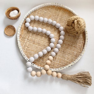 Könnte beinhalten: Eine weiße Holzperlenkette mit einem natürlichen Jute-Quaste, die auf einem geflochtenen Weidentablett hängt. Die Girlande besteht aus runden Perlen und ist mit einem natürlichen Garn zusammengefügt.
