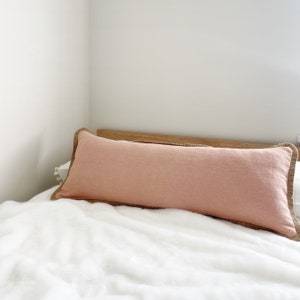 Puede incluir: Una almohada larga de color rosa claro con ribete tejido marrón sobre una cama blanca.