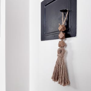 Może przedstawiać: Brązowo-beżowa frędzla z drewnianymi koralikami wisząca na czarnym haczyku na białej ścianie.