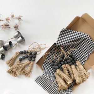 Peut inclure: Un ensemble de trois pompons noirs perlés avec une frange en jute, chacun d'une longueur différente. Les pompons sont présentés sur un tissu à carreaux noir et blanc.