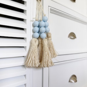 Peut inclure: Un ensemble de trois glands en bois de couleur bleu clair avec une frange en fibre naturelle. Les glands sont suspendus à un placard blanc.