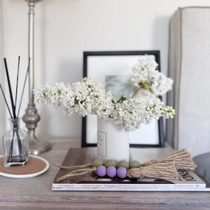 May include: A white ceramic vase holds a bouquet of white flowers. A decorative wooden bead garland with green and purple beads and jute tassels rests on a magazine. A reed diffuser and a framed picture are also visible.