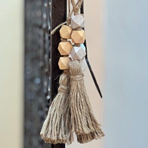 Pode incluir: Um conjunto de contas de madeira em tons bege e cinza com um tassel de juta natural. As contas são de forma geométrica e são enfiadas em um cordão.