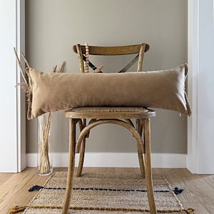 Puede incluir: Un cojín lumbar de color marrón claro con borde de flecos se coloca sobre una silla de madera con asiento tejido. La silla está frente a una pared de color beige claro.