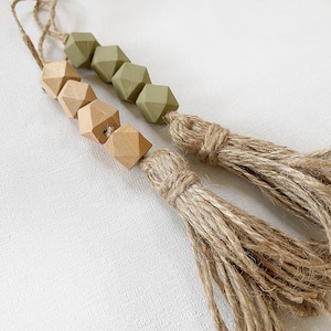 Op de afbeelding: Een houten kralenslinger met groene en natuurlijke houten kralen, geaccentueerd met twee jute kwastjes.