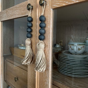 Peut inclure: Deux glands noirs à franges brunes suspendus aux boutons de cabinet. L'armoire est en bois clair avec une façade en verre.