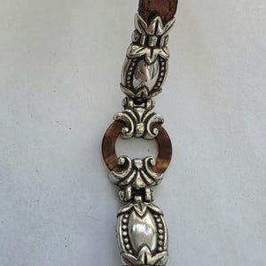 May include: A bracelet featuring a brown leather strap with a crocodile-embossed texture, accented by polished silver-tone metal links. The design includes two elongated, ornate silver-tone beads and a central circular brown resin ring.
