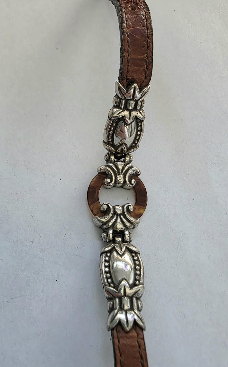 May include: A bracelet featuring a brown leather strap with a crocodile-embossed texture, accented by polished silver-tone metal links. The design includes two elongated, ornate silver-tone beads and a central circular brown resin ring.