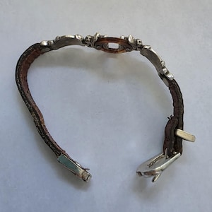 May include: A bracelet featuring a brown textured leather strap with silver-toned metal accents. The center piece is a circular brown resin element framed by silver metal. The bracelet includes a silver-toned metal buckle closure mechanism.