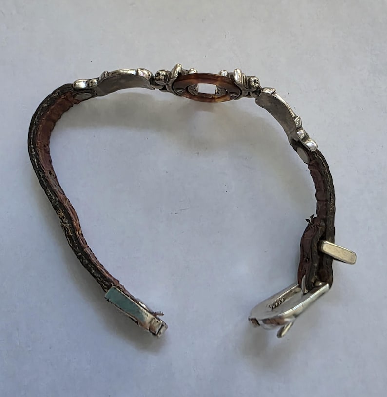 May include: A bracelet featuring a brown textured leather strap with silver-toned metal accents. The center piece is a circular brown resin element framed by silver metal. The bracelet includes a silver-toned metal buckle closure mechanism.