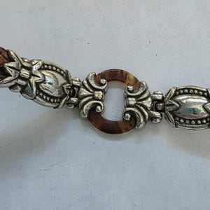 May include: A brown leather belt featuring two ornate silver-toned metal accents with intricate embossed detailing. The center of the belt is connected by a circular, tortoiseshell-patterned resin ring flanked by two silver-toned metal filigree pieces.