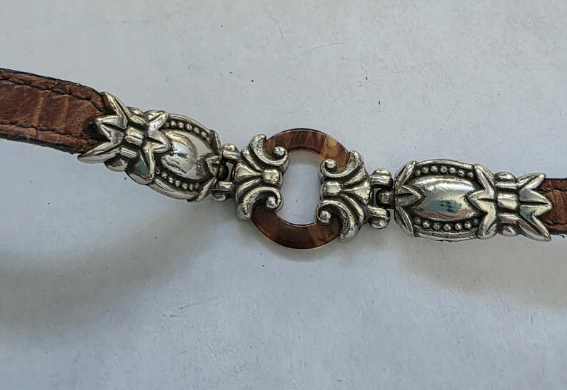 May include: A brown leather belt featuring two ornate silver-toned metal accents with intricate embossed detailing. The center of the belt is connected by a circular, tortoiseshell-patterned resin ring flanked by two silver-toned metal filigree pieces.