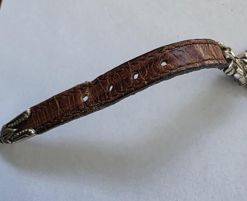 May include: A brown leather watch strap with a textured, crocodile-embossed pattern. The strap features four circular adjustment holes and is finished with silver-toned metal hardware at the tip and the attachment point to the watch case.