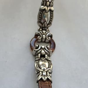 May include: A brown leather strap with a crocodile-embossed texture, featuring two ornate silver-toned metal decorative links connected by a small, circular, translucent amber-colored ring. The metal components have intricate floral-inspired detailing.