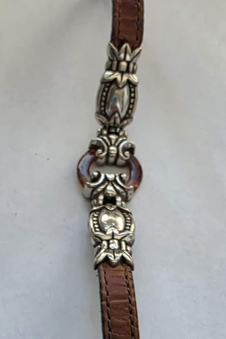 May include: A brown leather strap with a crocodile-embossed texture, featuring two ornate silver-toned metal decorative links connected by a small, circular, translucent amber-colored ring. The metal components have intricate floral-inspired detailing.