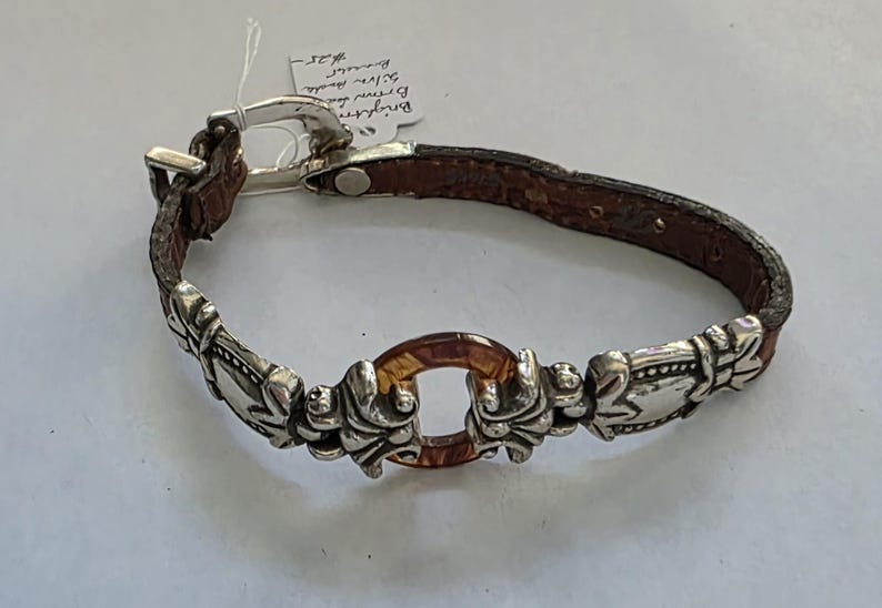 May include: A brown leather bracelet featuring silver-toned metal accents and a central amber-colored resin ring. The bracelet includes a silver-toned metal buckle closure and a white paper tag with handwritten text attached to the buckle.