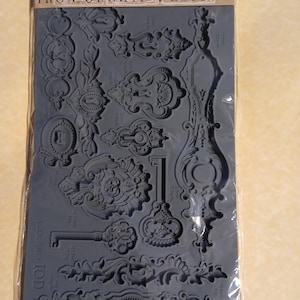 Op de afbeelding: Een grijze siliconen mal met sierlijke slot- en sleutelontwerpen. De mal bevat verschillende vormen en maten van sleutels, sloten en decoratieve elementen. De verpakking leest "IRON ORCHID DESIGNS MOULD 'LOCK & KEY'".