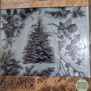 Könnte beinhalten: Klarstempel-Set mit Stechpalmen- und Kiefernmotiven. Das Set enthält botanische Elemente wie Kiefernnadeln, Tannenzapfen und einen detaillierten Weihnachtsbaum. Die Verpackung trägt den Text "Iron Orchid Designs" und "Holly & Pine".
