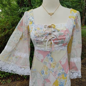 May include: A vintage patchwork dress with a square neckline and long, bell sleeves. The dress features a mix of floral and geometric patterns in pastel colors, with white lace trim and a ribbon tie at the bodice. A gold necklace with a pendant is also visible.