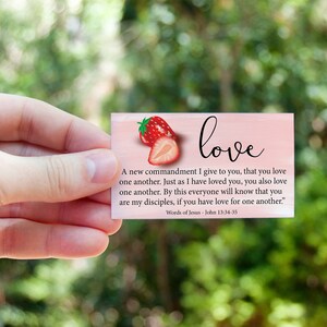 K&ouml;nnte beinhalten: Eine rosa Karte mit einer Erdbeerillustration und dem Wort "love" in kursiver Schrift. Die Karte zeigt ein Zitat aus der Bibel, Johannes 13,34-35, &uuml;ber die Liebe zueinander. "Ein neues Gebot gebe ich euch: Liebt einander! Wie ich euch geliebt habe, so sollt auch ihr einander lieben. Daran werden alle erkennen, dass ihr meine J&uuml;nger seid, wenn ihr Liebe untereinander habt." Worte Jesu - Johannes 13,34-35