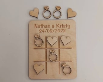 Könnte beinhalten: Ein Tic-Tac-Toe-Spiel aus Holz mit den Namen Nathan & Kristy und dem Datum 24.09.2022. Die Spielsteine sind Herzen und Ringe.