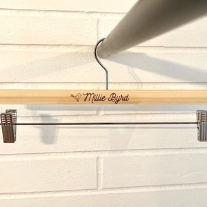 May include: Wooden hanger with a metal bar and clips. The hanger has the name "Millie Byrd" engraved on it.