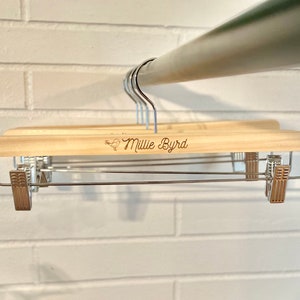 May include: Wooden hanger with a metal bar and clips. The hanger is engraved with the name "Millie Byrd".