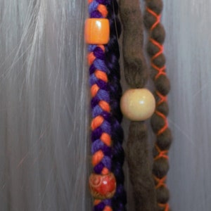 May include: A close-up of a hair extension with orange, purple, and brown strands. The extension is braided and has wooden beads.