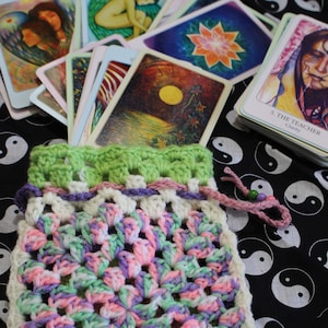 May include: A crocheted bag with a green, pink, and white pattern. The bag is filled with a deck of tarot cards. The cards feature colorful illustrations and the text "5. The Teacher Clarity".