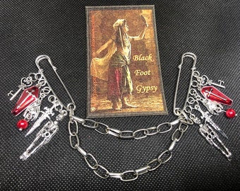 May include: Two silver safety pin brooches connected by a chain. Each brooch features a red coffin charm, a skeleton charm, a key charm, a dagger charm, and a red bead charm.