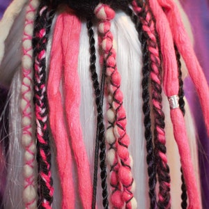 Könnte beinhalten: Nahaufnahme einer Sammlung von Dreadlocks in verschiedenen Farben. Die Dreadlocks sind hauptsächlich rosa, schwarz und cremefarben, mit silbernen Akzenten. Einige Dreadlocks sind geflochten, andere sind einfarbig. Der Hintergrund ist ein verschwommenes Lila.