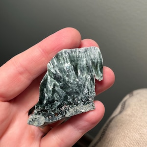 May include: A piece of raw seraphinite gemstone, held in a hand. The stone has a dark green color with a feathery, fibrous texture and a silvery sheen. The bottom edge has a rough, gray appearance.