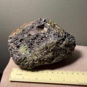 May include: A porous volcanic rock with a dark grey and olive green textured surface, positioned above a yellow plastic ruler that shows the specimen measures approximately 5.5 inches in length along its base.