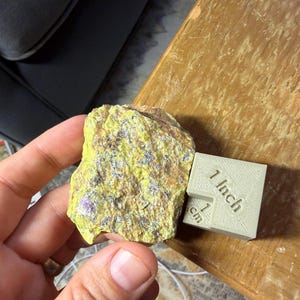 May include: A rough, irregular mineral specimen featuring a vibrant neon yellow surface with metallic silver inclusions. Next to the mineral is a beige 3D-printed scale cube clearly marked with the text 1 Inch and 1 cm for size reference.