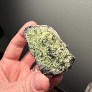 May include: A hand holding a rough-hewn rock specimen. The rock has a dark gray exterior and a vibrant, light green, crystalline interior. Small dark flecks are visible within the green matrix. The specimen is a natural mineral formation.