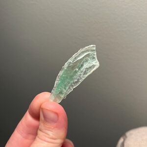 May include: A translucent, irregularly shaped piece of green mineral, held in a hand. The mineral has a textured surface with visible internal structures. The color is a pale green, with some areas appearing more transparent than others.