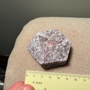 May include: A raw, hexagonal-shaped pink and grey mineral specimen with a crystalline, textured surface. A yellow plastic ruler is positioned along the bottom edge, indicating the stone measures approximately 1.75 inches in width.