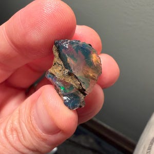May include: A raw, translucent opal gemstone featuring a play of color with flashes of blue, green, and orange. The stone has an irregular, jagged shape with a rough, textured brown matrix vein running diagonally across the surface of the crystal.